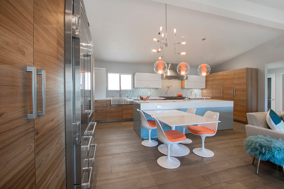 This is an example of a large midcentury l-shaped eat-in kitchen in Los Angeles with a farmhouse sink, flat-panel cabinets, medium wood cabinets, quartzite benchtops, blue splashback, porcelain splashback, stainless steel appliances, laminate floors, with island, brown floor and white benchtop.