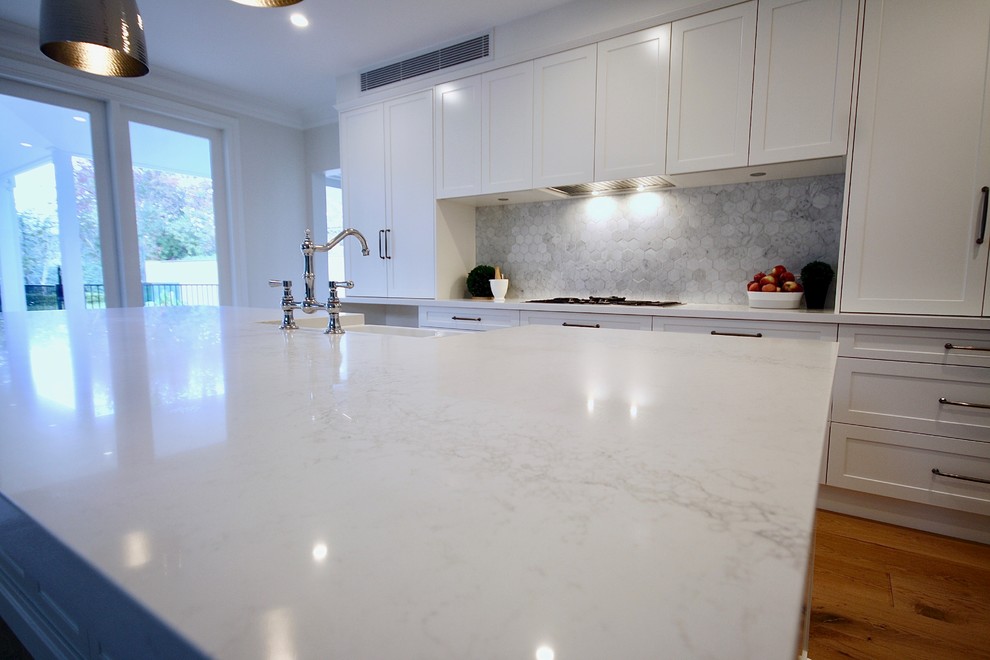 Inspiration for a large transitional l-shaped eat-in kitchen in Sydney with a farmhouse sink, shaker cabinets, white cabinets, quartz benchtops, grey splashback, ceramic splashback, black appliances, medium hardwood floors, with island, brown floor and white benchtop.