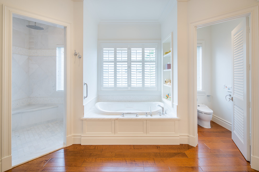 Photo of an expansive transitional master bathroom in Miami with an undermount sink, furniture-like cabinets, white cabinets, marble benchtops, a drop-in tub, an open shower, a one-piece toilet, white tile, stone slab, white walls and medium hardwood floors.