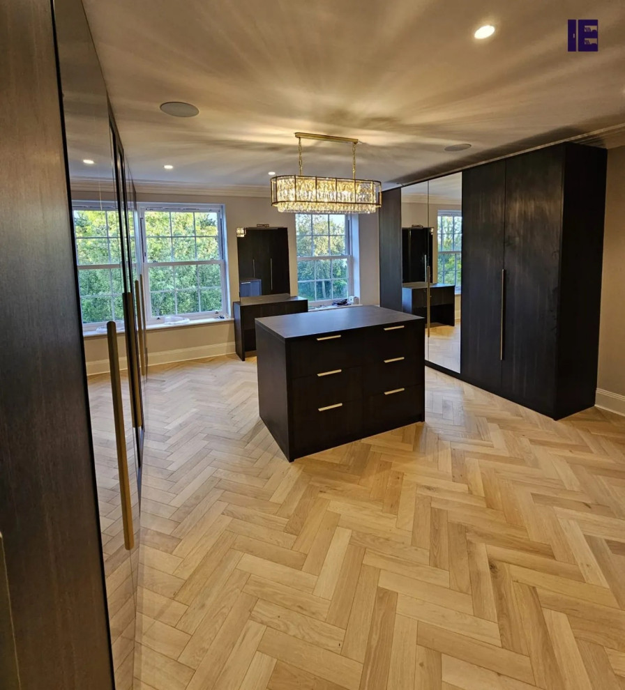 Large modern walk-in wardrobe in London with glass-front cabinets, dark wood cabinets and plywood floors.