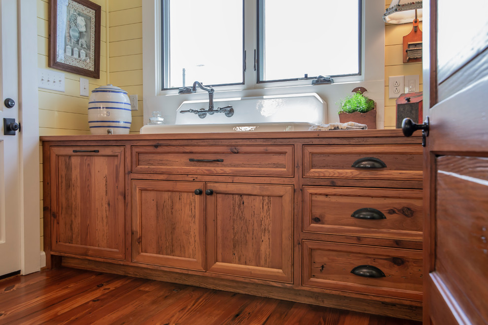 Design ideas for a mid-sized country single-wall utility room in Charlotte with a farmhouse sink, beaded inset cabinets, medium wood cabinets, wood benchtops, yellow walls, medium hardwood floors and a concealed washer and dryer.