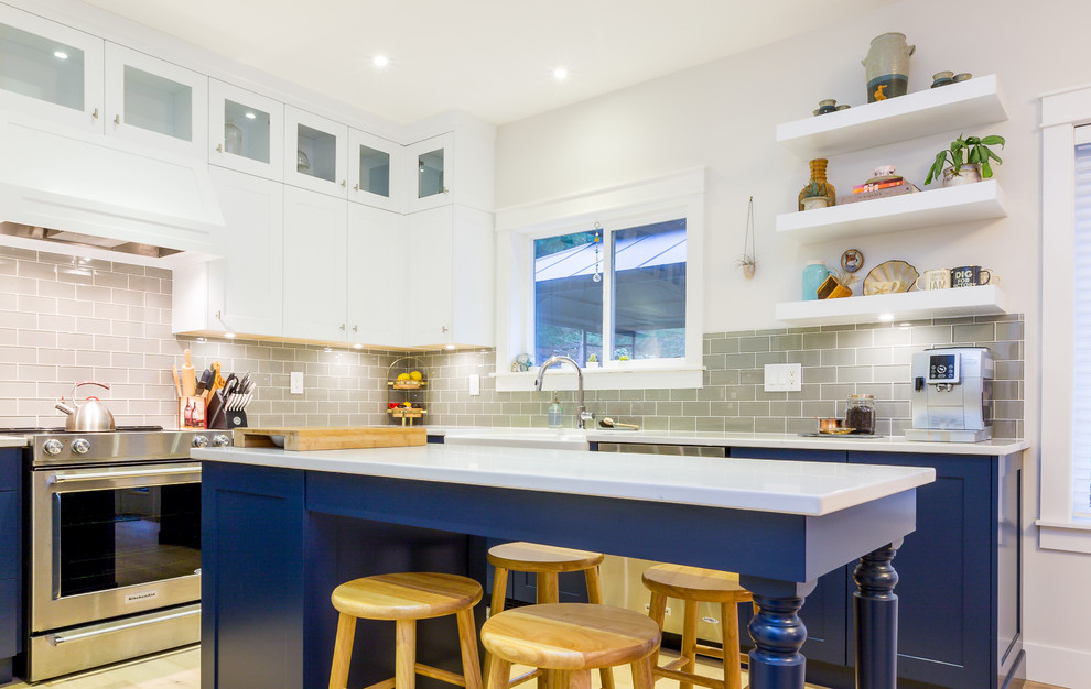 Large transitional u-shaped light wood floor open concept kitchen photo with a farmhouse sink, shaker cabinets, blue cabinets, quartz countertops, gray backsplash, glass tile backsplash, stainless steel appliances, an island and white countertops