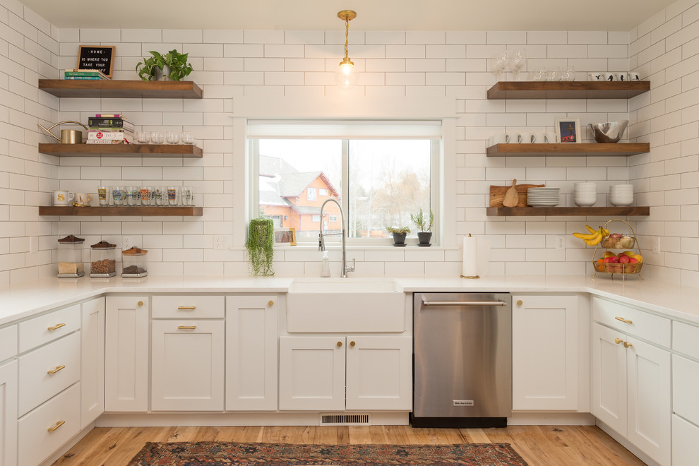 Photo of a transitional u-shaped kitchen in Other with a farmhouse sink, shaker cabinets, white cabinets, white splashback, stainless steel appliances, medium hardwood floors, brown floor and white benchtop.