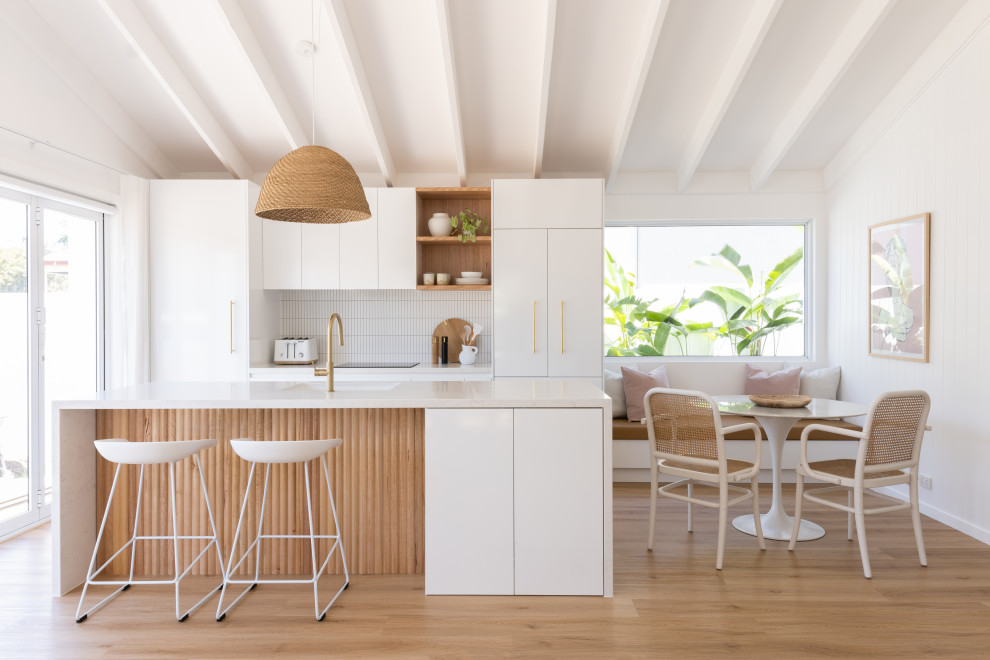 Bild på ett maritimt vit vitt kök, med vitt stänkskydd, stänkskydd i keramik, en undermonterad diskho, släta luckor, vita skåp, integrerade vitvaror, mellanmörkt trägolv, en köksö och brunt golv