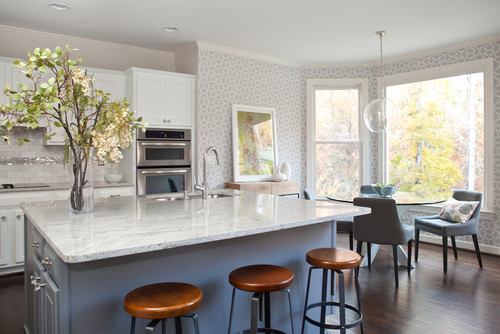Modern kitchen cabinet design with white shaker cabinets, a large quartz island, wooden bar stools, and a bright breakfast nook overlooking the trees.