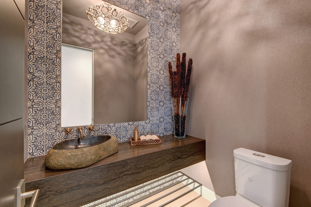 Inspiration for a mid-sized eclectic powder room in Other with brown cabinets, a two-piece toilet, blue tile, terra-cotta tile, grey walls, porcelain floors, a vessel sink, limestone benchtops, grey floor, brown benchtops, a floating vanity, wallpaper and wallpaper.