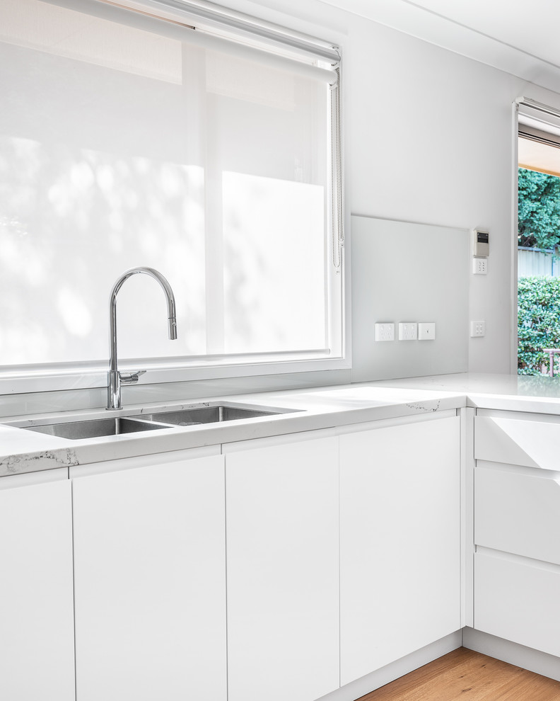 Mid-sized contemporary u-shaped eat-in kitchen in Sydney with a double-bowl sink, raised-panel cabinets, white cabinets, solid surface benchtops, white splashback, glass sheet splashback, stainless steel appliances, light hardwood floors, no island, brown floor and white benchtop.