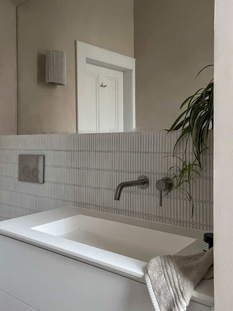 Foto på ett litet minimalistiskt vit badrum för barn, med släta luckor, vita skåp, ett platsbyggt badkar, en dusch/badkar-kombination, en vägghängd toalettstol, vit kakel, mosaik, beige väggar, ett väggmonterat handfat, bänkskiva i akrylsten, beiget golv och med dusch som är öppen