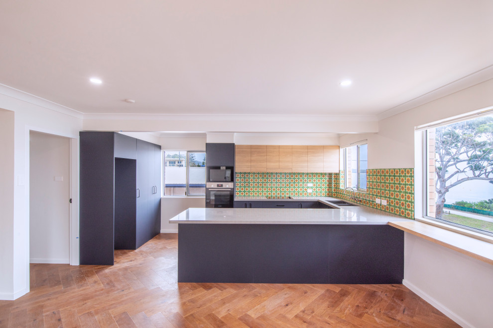 Inspiration for a small midcentury u-shaped kitchen pantry in Central Coast with a drop-in sink, blue cabinets, multi-coloured splashback, black appliances, light hardwood floors, with island and grey benchtop.