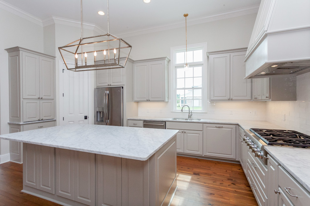 Inspiration for a mid-sized traditional l-shaped open plan kitchen in New Orleans with a drop-in sink, raised-panel cabinets, grey cabinets, marble benchtops, white splashback, ceramic splashback, stainless steel appliances, medium hardwood floors and with island.