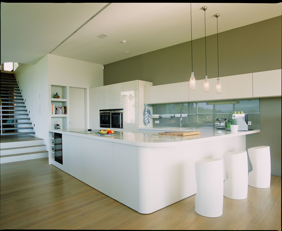 Photo of a large contemporary galley kitchen in Other with a double-bowl sink, flat-panel cabinets, white cabinets, brown splashback, glass sheet splashback, black appliances, medium hardwood floors and with island.
