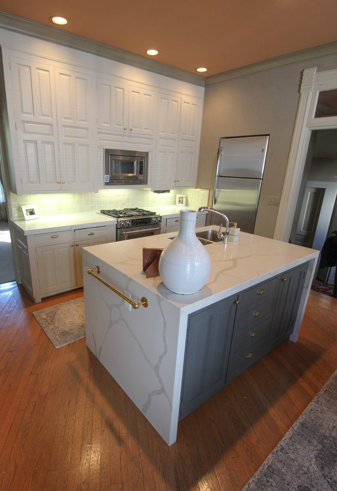 Transitional medium tone wood floor and brown floor kitchen photo with an undermount sink, raised-panel cabinets, white cabinets, quartz countertops, white backsplash, ceramic backsplash, stainless steel appliances, an island and white countertops