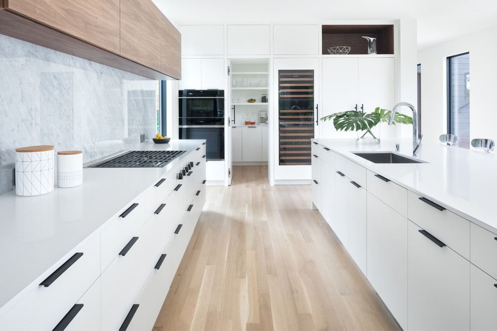 Photo of a contemporary kitchen in Minneapolis with an undermount sink, flat-panel cabinets, white cabinets, grey splashback, stone slab splashback, black appliances, light hardwood floors, with island, beige floor and white benchtop.