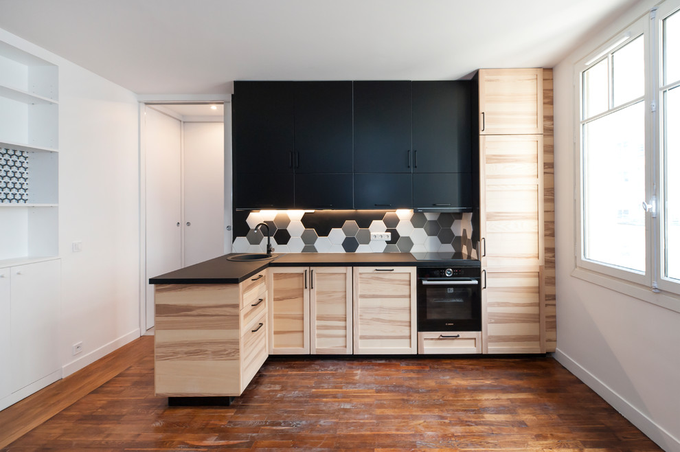Design ideas for a mid-sized contemporary l-shaped kitchen in Paris with a drop-in sink, shaker cabinets, light wood cabinets, multi-coloured splashback, black appliances, medium hardwood floors, a peninsula, brown floor and black benchtop.