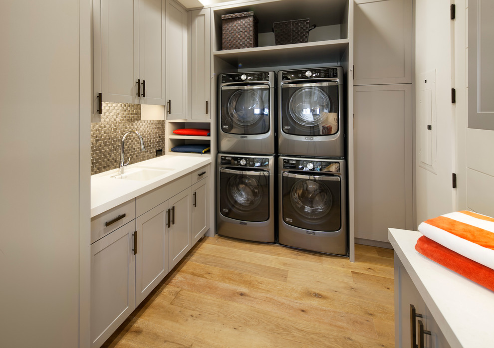 Design ideas for a mid-sized mediterranean u-shaped dedicated laundry room in Santa Barbara with an undermount sink, shaker cabinets, grey cabinets, solid surface benchtops, white walls, light hardwood floors, a stacked washer and dryer and white benchtop.