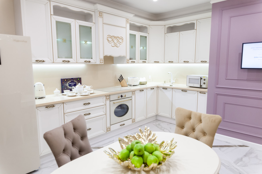Mid-sized transitional l-shaped eat-in kitchen in Other with an integrated sink, glass-front cabinets, white cabinets, solid surface benchtops, beige splashback, stone slab splashback, white appliances, porcelain floors, no island, white floor and beige benchtop.