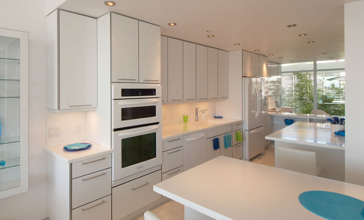 Design ideas for a large modern galley open plan kitchen in Vancouver with an undermount sink, flat-panel cabinets, white cabinets, quartzite benchtops, white splashback, stone slab splashback, white appliances, travertine floors, with island, beige floor and white benchtop.