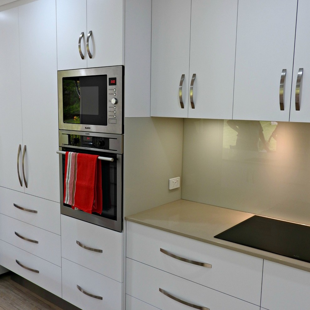 This is an example of a mid-sized contemporary galley kitchen pantry in Brisbane with a single-bowl sink, flat-panel cabinets, white cabinets, quartz benchtops, beige splashback, glass sheet splashback, stainless steel appliances, vinyl floors, a peninsula and brown floor.