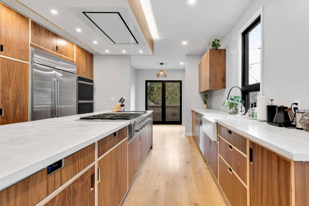 Large contemporary galley kitchen in Baltimore with a farmhouse sink, flat-panel cabinets, dark wood cabinets, concrete benchtops, stainless steel appliances, light hardwood floors, with island and white benchtop.