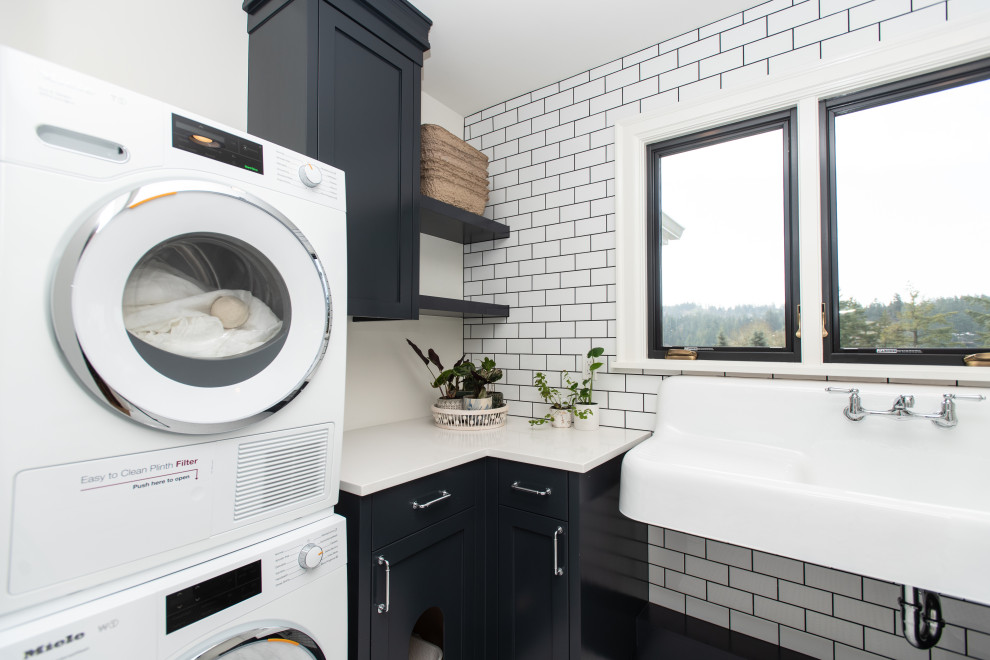 Photo of a mid-sized traditional u-shaped laundry room in Portland with shaker cabinets, black cabinets, an utility sink, white splashback, stone slab splashback, white walls, a stacked washer and dryer, white floor and white benchtop.