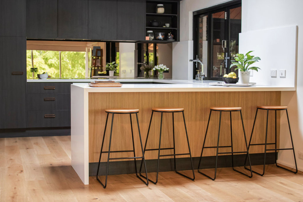 Mid-sized midcentury u-shaped open plan kitchen in Adelaide with a double-bowl sink, flat-panel cabinets, black cabinets, quartz benchtops, brown splashback, glass sheet splashback, stainless steel appliances, medium hardwood floors, a peninsula, brown floor, white benchtop and exposed beam.