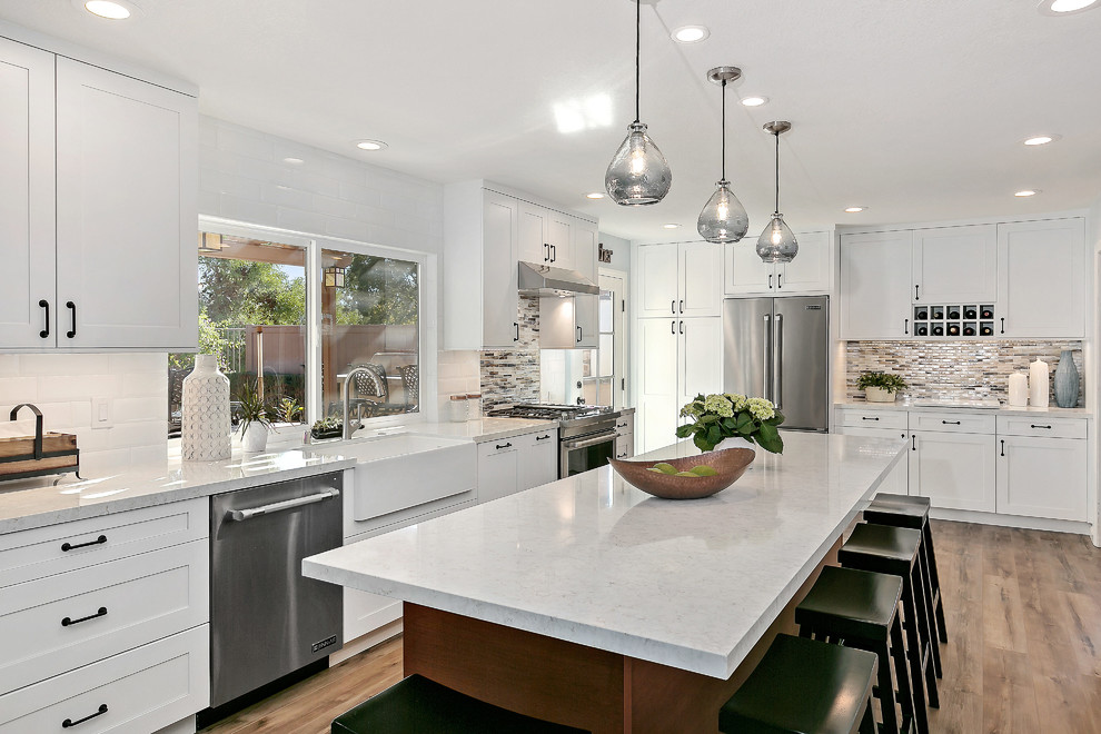 Inspiration for a large transitional l-shaped open plan kitchen in Tampa with a farmhouse sink, shaker cabinets, white cabinets, quartz benchtops, white splashback, porcelain splashback, stainless steel appliances, vinyl floors, with island, brown floor and white benchtop.