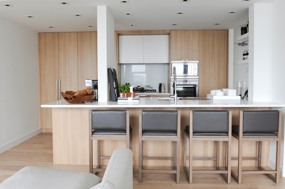 This is an example of a contemporary galley open plan kitchen in Vancouver with flat-panel cabinets, light wood cabinets, white splashback, glass sheet splashback and panelled appliances.