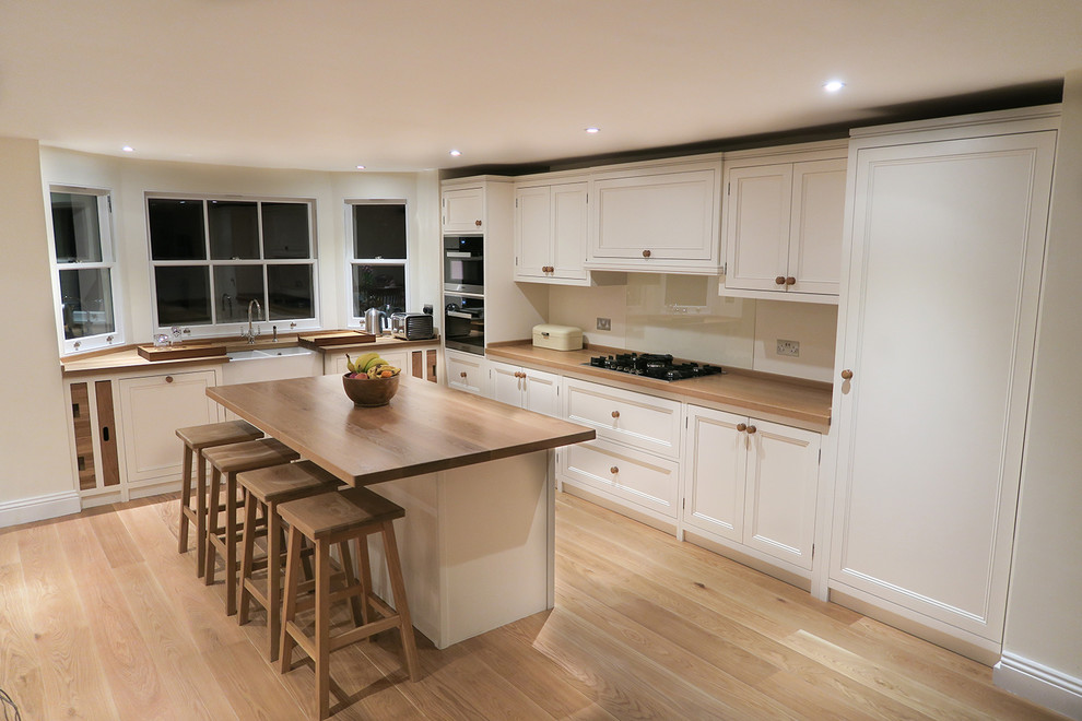 This is an example of a large transitional l-shaped kitchen in London with an undermount sink, recessed-panel cabinets, white cabinets, wood benchtops, stainless steel appliances, medium hardwood floors and with island.