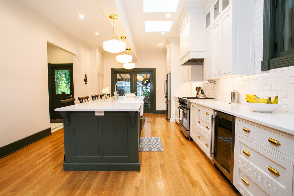 Example of a transitional galley medium tone wood floor and brown floor kitchen design in Indianapolis with a farmhouse sink, beaded inset cabinets, white cabinets, white backsplash, subway tile backsplash, stainless steel appliances, an island and white countertops