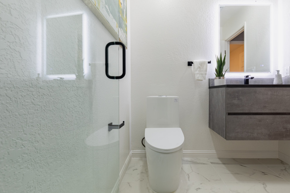 Inspiration for a mid-sized contemporary 3/4 bathroom in San Diego with grey cabinets, ceramic tile, white walls, ceramic floors, engineered quartz benchtops, white floor, flat-panel cabinets, a corner tub, gray tile, a hinged shower door, black benchtops, a single vanity, a floating vanity, a one-piece toilet, an integrated sink, brick walls and an alcove shower.