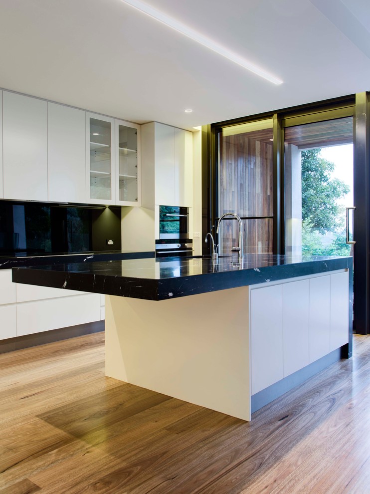 Design ideas for a large contemporary galley open plan kitchen in Melbourne with an undermount sink, flat-panel cabinets, white cabinets, marble benchtops, black splashback, glass sheet splashback, stainless steel appliances, light hardwood floors and multiple islands.