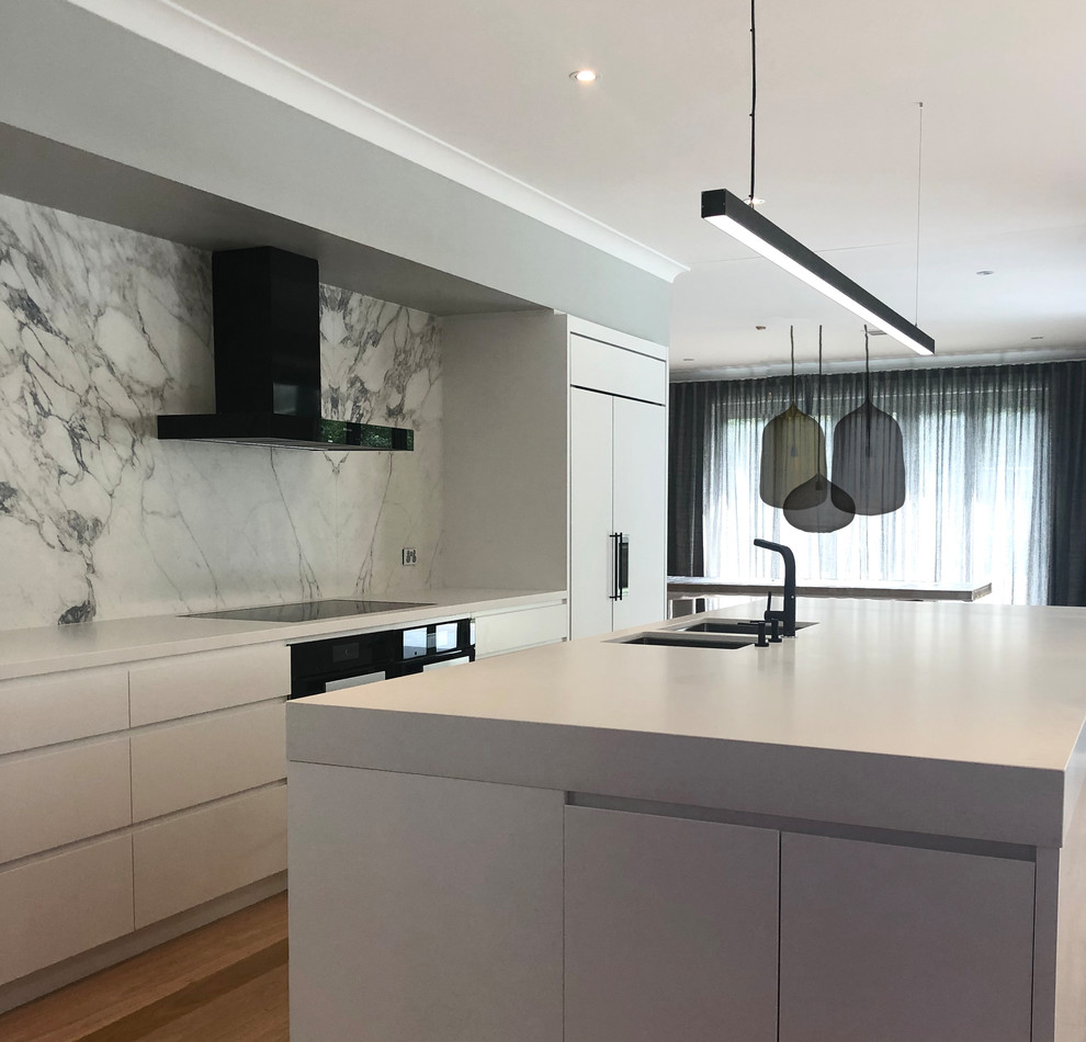 Large contemporary galley kitchen in Canberra - Queanbeyan with a drop-in sink, flat-panel cabinets, white cabinets, quartz benchtops, multi-coloured splashback, marble splashback, panelled appliances, light hardwood floors, with island, brown floor and white benchtop.