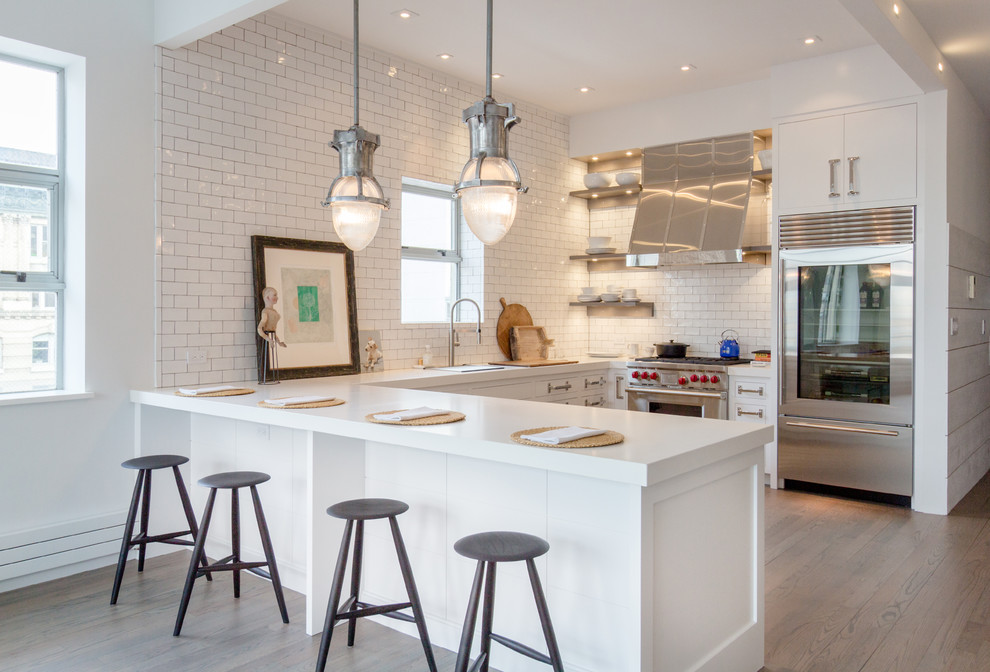 Photo of a mid-sized industrial u-shaped open plan kitchen in New York with white cabinets, quartz benchtops, white splashback, subway tile splashback, stainless steel appliances, light hardwood floors, a peninsula and flat-panel cabinets.