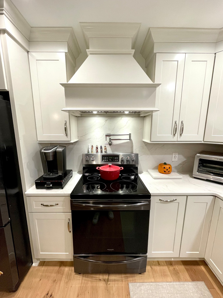 Transitional l-shaped light wood floor and beige floor eat-in kitchen photo with a farmhouse sink, shaker cabinets, white cabinets, quartz countertops, white backsplash, quartz backsplash, black appliances, an island and white countertops