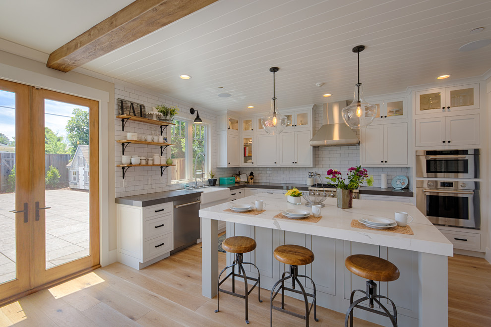 This is an example of a large country l-shaped separate kitchen in San Francisco with a farmhouse sink, shaker cabinets, white cabinets, white splashback, subway tile splashback, stainless steel appliances, medium hardwood floors, with island, brown floor and quartzite benchtops.
