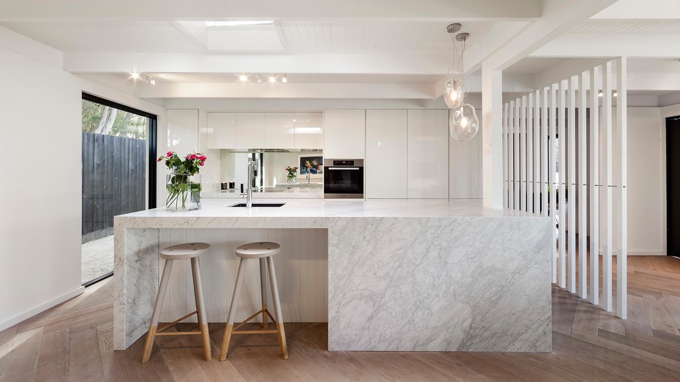 Contemporary galley open plan kitchen in Melbourne with an undermount sink, flat-panel cabinets, white cabinets, metallic splashback, mirror splashback, stainless steel appliances, light hardwood floors and with island.