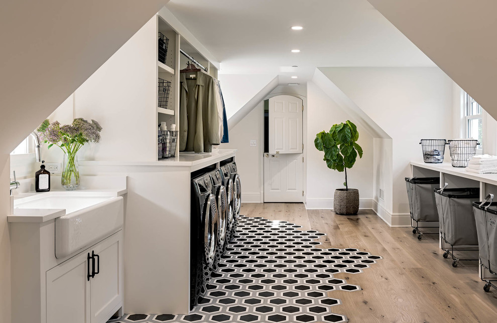 Photo of a mid-sized country galley dedicated laundry room in New York with a farmhouse sink, shaker cabinets, white cabinets, white walls, medium hardwood floors, a side-by-side washer and dryer and brown floor.