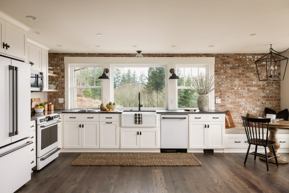 This is an example of a mid-sized country l-shaped open plan kitchen in Portland with a farmhouse sink, shaker cabinets, white cabinets, quartz benchtops, brick splashback, white appliances, dark hardwood floors, no island and black benchtop.
