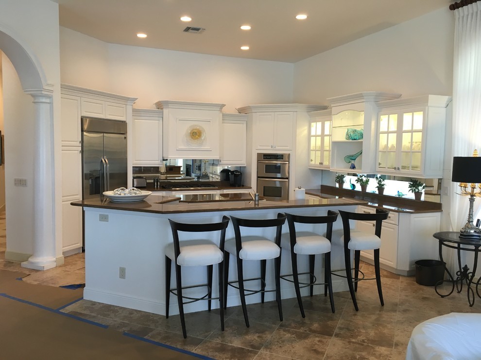 Mid-sized contemporary l-shaped open plan kitchen in Miami with mirror splashback, raised-panel cabinets, white cabinets, granite benchtops, stainless steel appliances, ceramic floors, multiple islands, brown floor and brown benchtop.