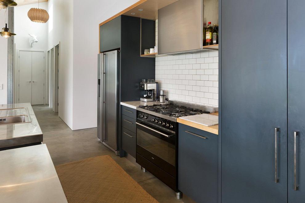 Photo of a large contemporary single-wall open plan kitchen in Auckland with a drop-in sink, flat-panel cabinets, blue cabinets, stainless steel benchtops, subway tile splashback, stainless steel appliances, concrete floors, with island and grey floor.