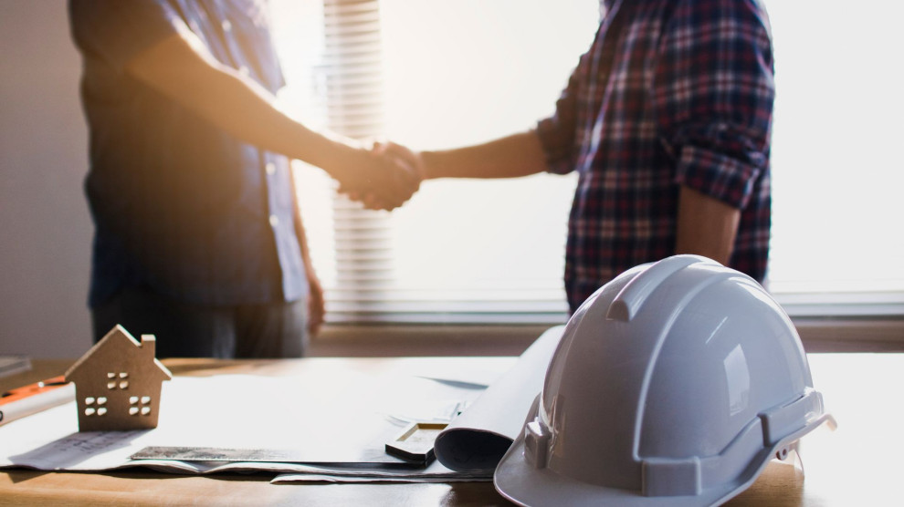 Two people shaking hands across a table with a hard hat, symbolizing trust and partnership in home remodeling.