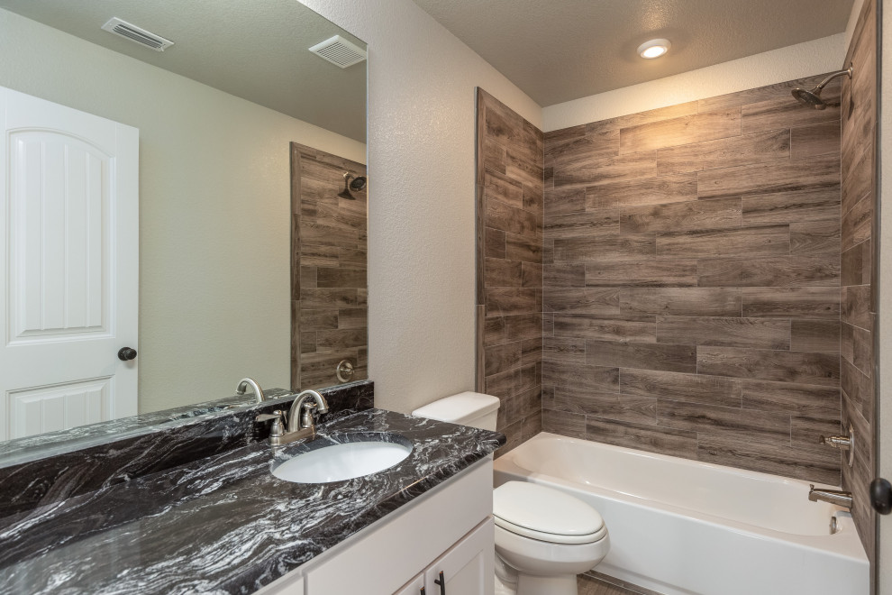 Mid-sized kids bathroom in Dallas with shaker cabinets, white cabinets, a drop-in tub, a curbless shower, a two-piece toilet, gray tile, wood-look tile, grey walls, vinyl floors, an undermount sink, granite benchtops, grey floor, a hinged shower door and black benchtops.