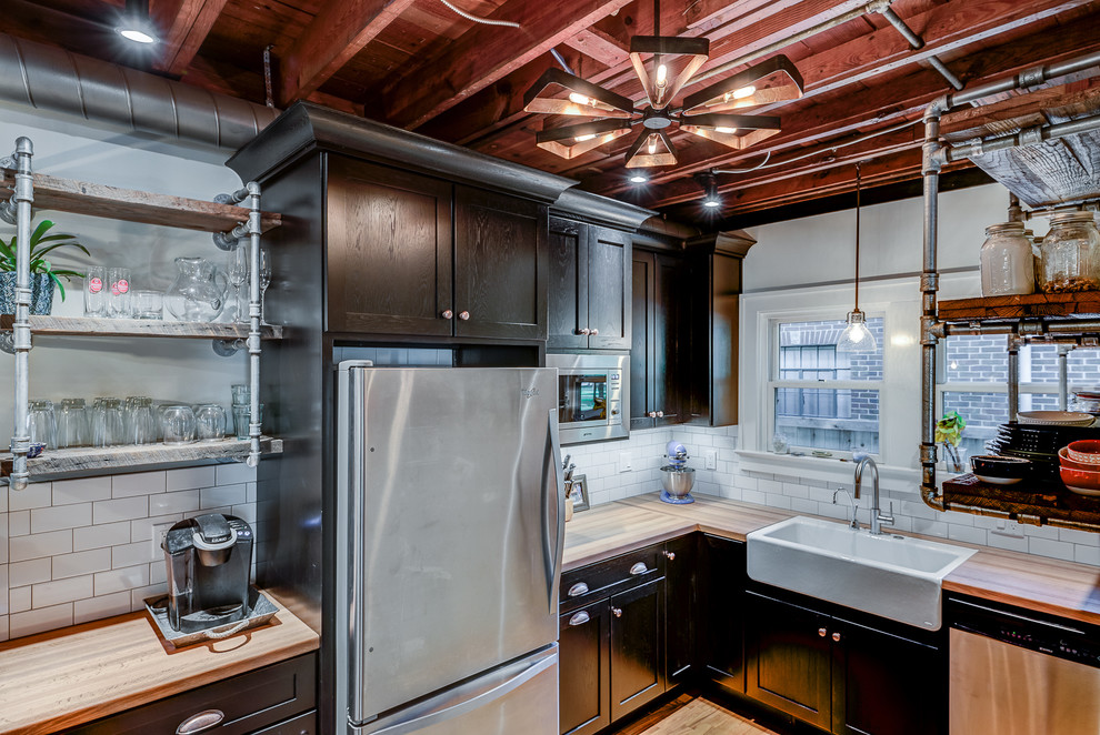 Photo of a small industrial u-shaped eat-in kitchen in Chicago with a farmhouse sink, shaker cabinets, black cabinets, wood benchtops, white splashback, stainless steel appliances, light hardwood floors, a peninsula and subway tile splashback.