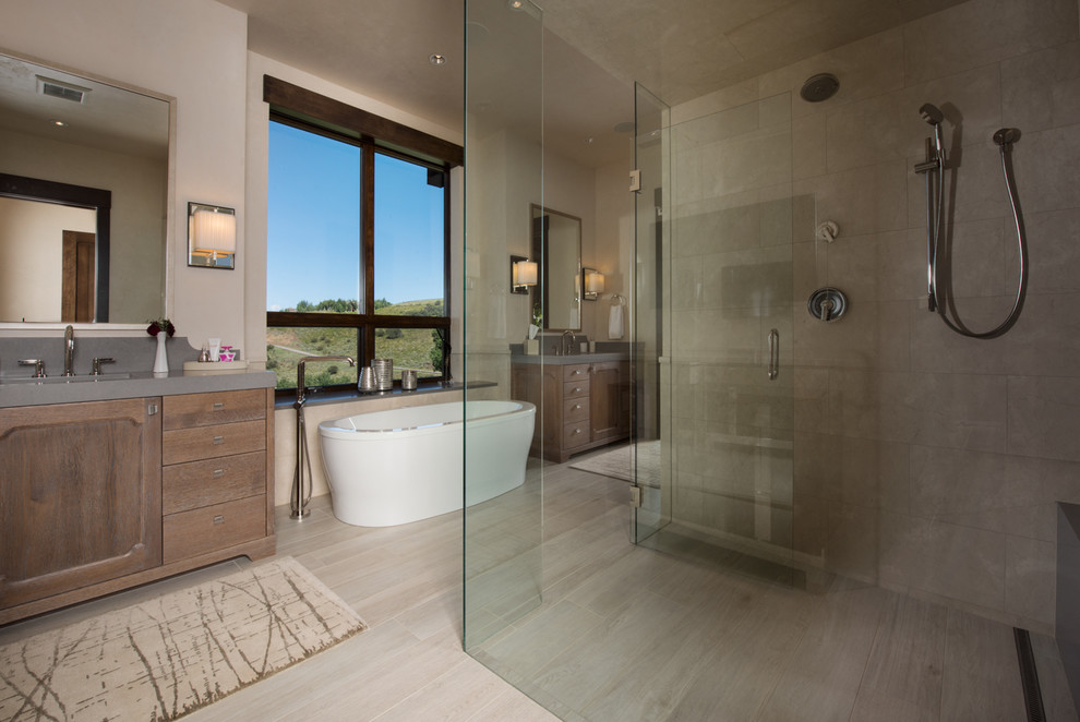 Photo of a large transitional master bathroom in Denver with recessed-panel cabinets, brown cabinets, a freestanding tub, a curbless shower, a one-piece toilet, beige tile, limestone, beige walls, porcelain floors, an undermount sink, engineered quartz benchtops, beige floor and a hinged shower door.