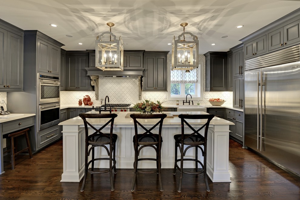 This is an example of a large transitional u-shaped kitchen in Minneapolis with grey cabinets, white splashback, stainless steel appliances, dark hardwood floors, with island, raised-panel cabinets, a farmhouse sink, solid surface benchtops, brown floor, white benchtop and ceramic splashback.