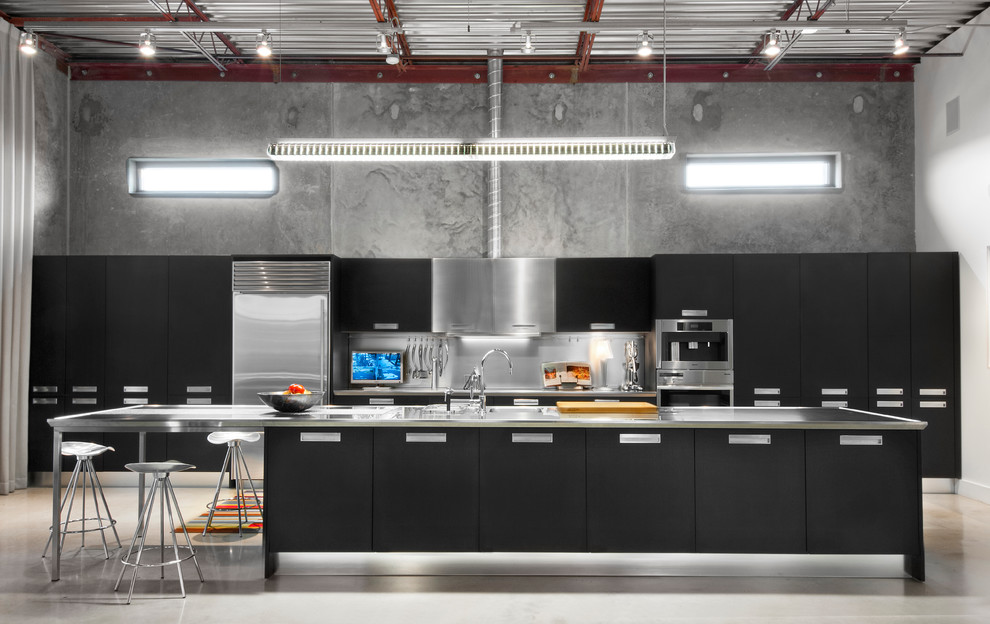 Design ideas for an industrial kitchen in Birmingham with an integrated sink, flat-panel cabinets, black cabinets, stainless steel benchtops, metallic splashback, stainless steel appliances, concrete floors and with island.