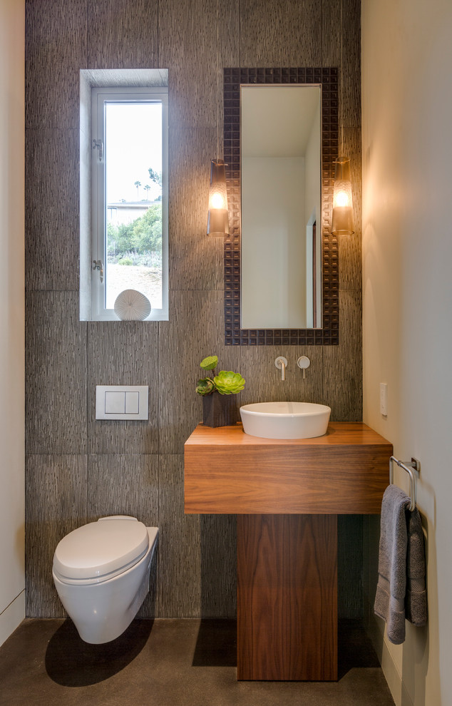 Contemporary powder room in San Francisco with a vessel sink, medium wood cabinets, a wall-mount toilet, gray tile, beige walls and concrete floors.