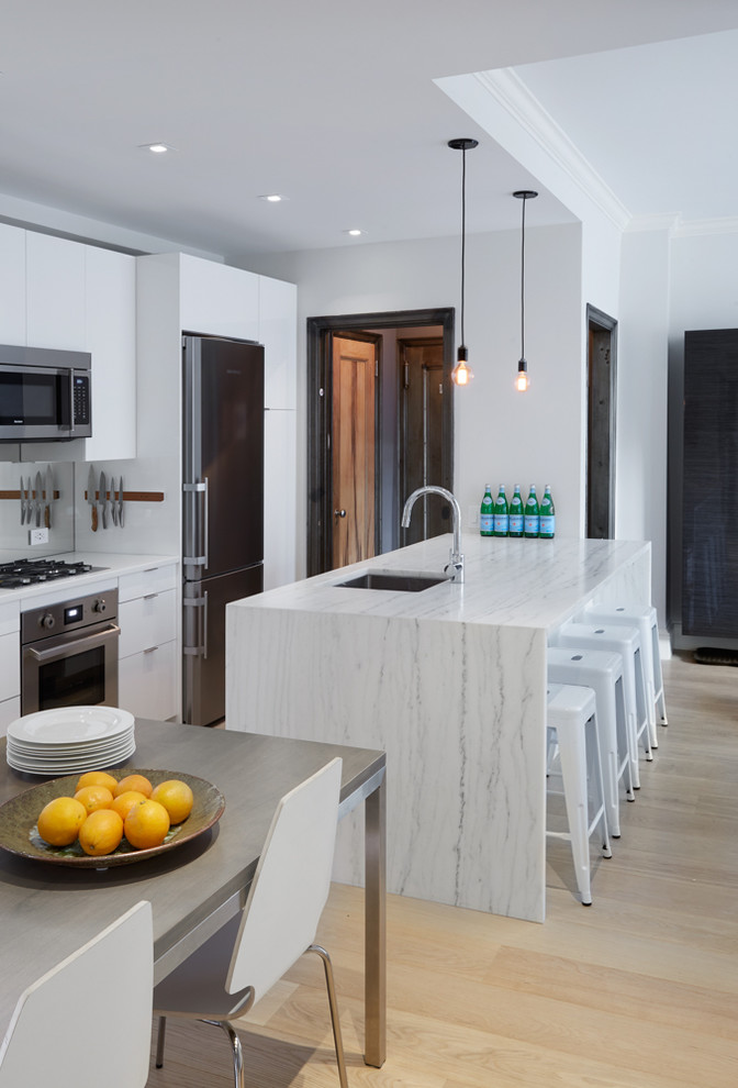This is an example of a small contemporary galley eat-in kitchen in New York with an undermount sink, flat-panel cabinets, white cabinets, marble benchtops, metallic splashback, mirror splashback, stainless steel appliances, light hardwood floors, a peninsula and beige floor.