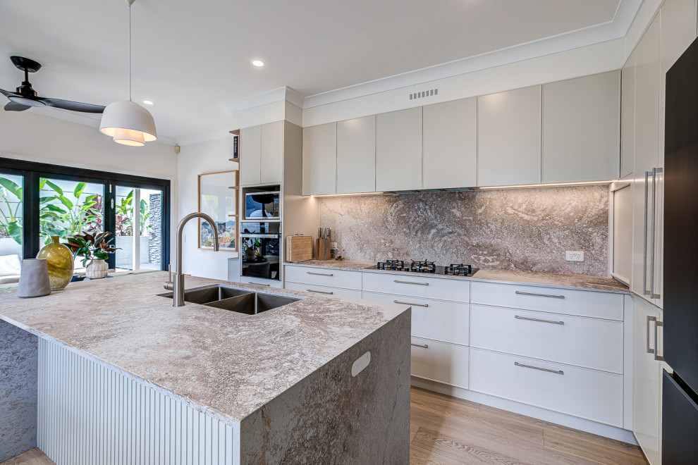 Design ideas for a large modern l-shaped open plan kitchen in Brisbane with a double-bowl sink, raised-panel cabinets, grey cabinets, quartz benchtops, engineered quartz splashback, black appliances, light hardwood floors, with island and brown floor.