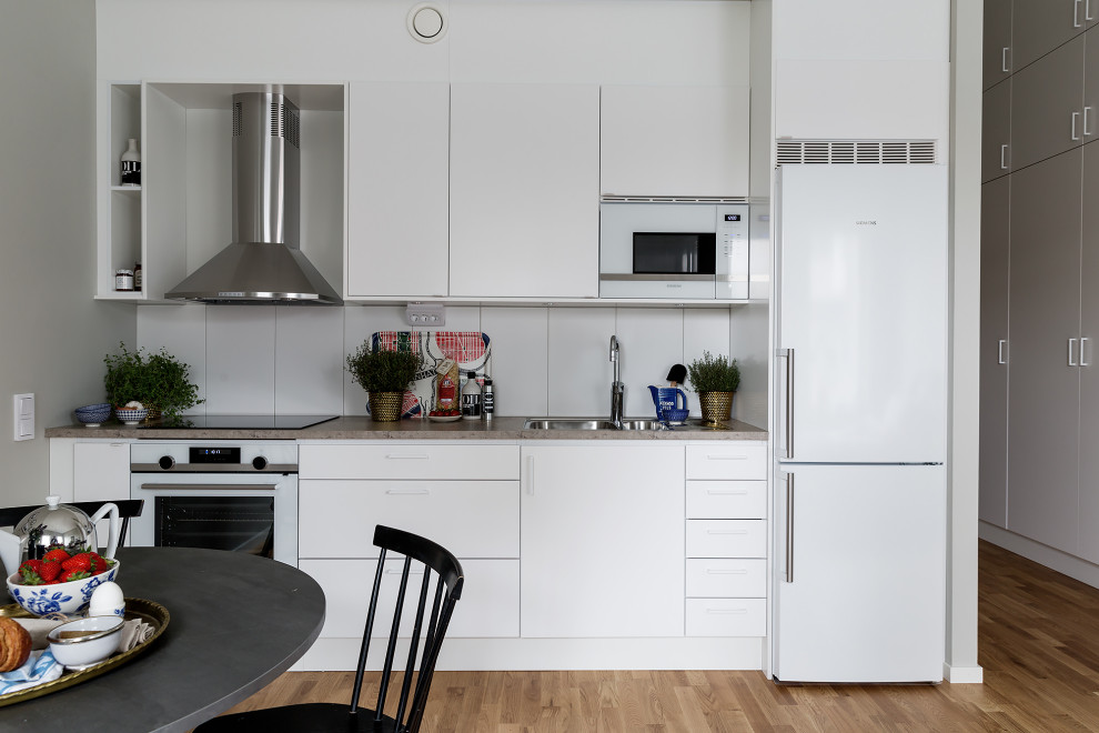 Photo of a contemporary single-wall open plan kitchen in Stockholm with an undermount sink, flat-panel cabinets, white cabinets, white appliances, medium hardwood floors, no island, brown floor and grey benchtop.